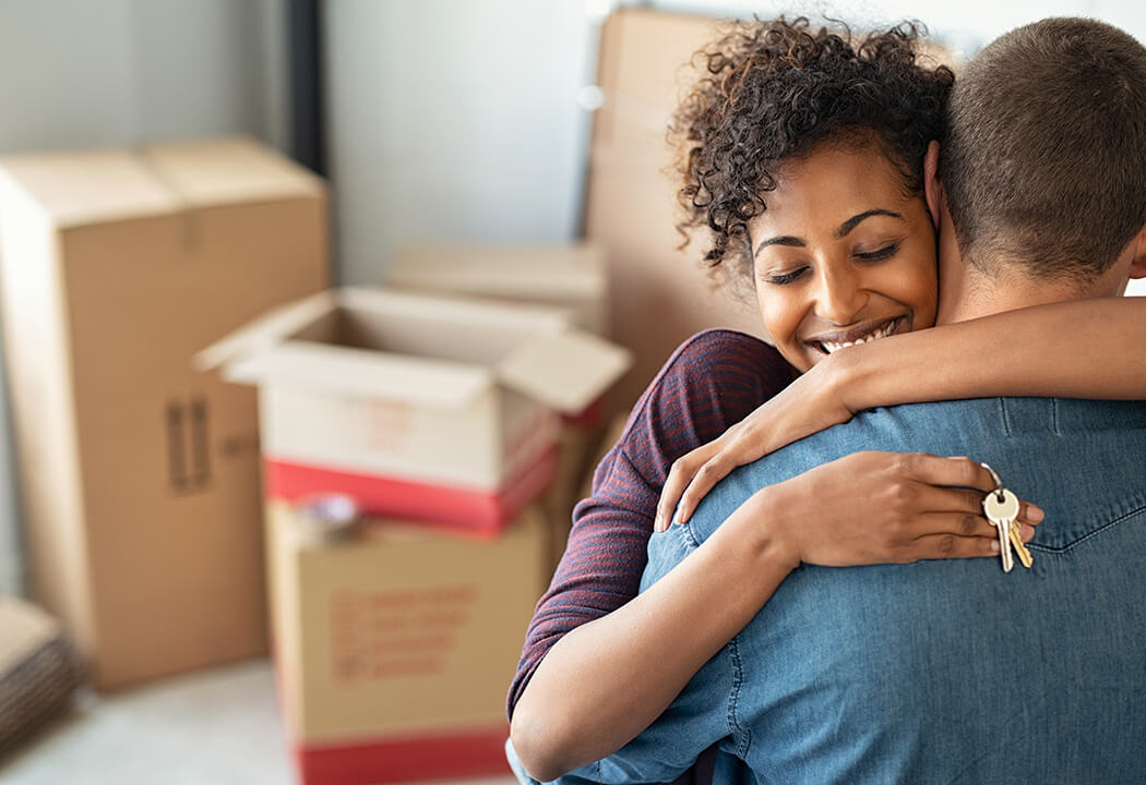 Man and woman embracing in a room with lots of moving boxes. The woman is holding house keys.